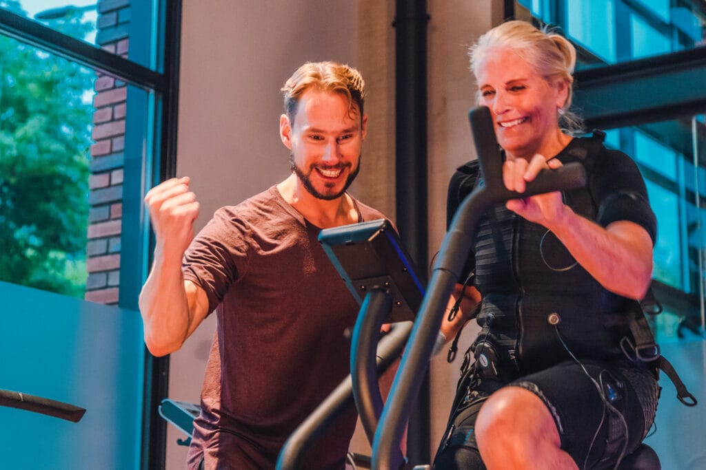 Een sporter die met EMS op een crosstrainer sport en ondersteund wordt door een personal trainer.